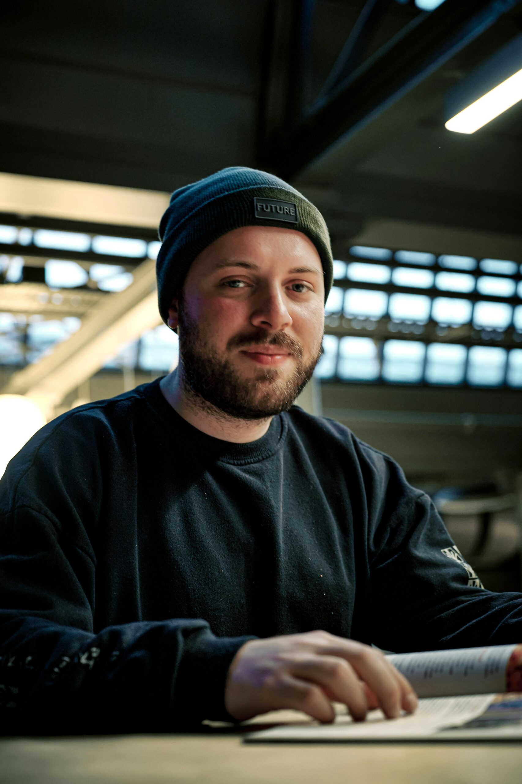 A man in a beanie reading a magazine indoors with a warm smile, capturing a relaxed atmosphere.